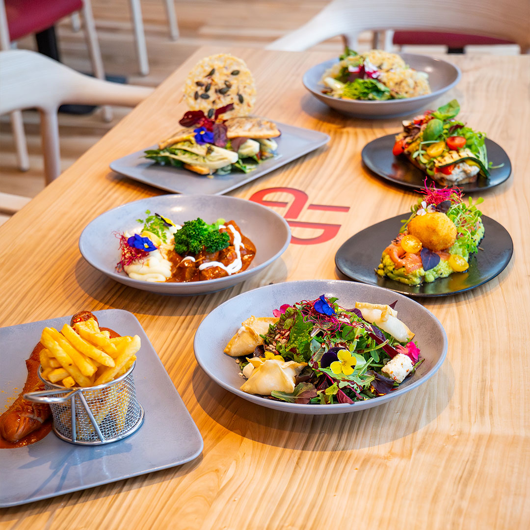 A wooden table laden with a variety of gourmet dishes, including salads with edible flowers, chips served in a basket, and a selection of colourful starters on grey plates, all arranged in a modern, brightly lit restaurant.