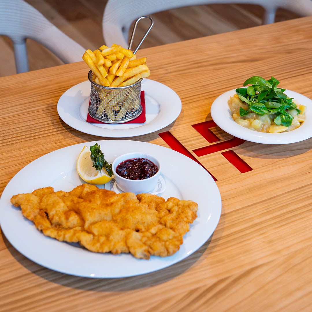 Ein Teller mit paniertem Schnitzel mit Zitrone, Beerensauce und Beilage, dazu Pommes frites in einem Metallkorb und ein Salat mit Kartoffeln und Grünzeug, alles auf einem Holztisch.