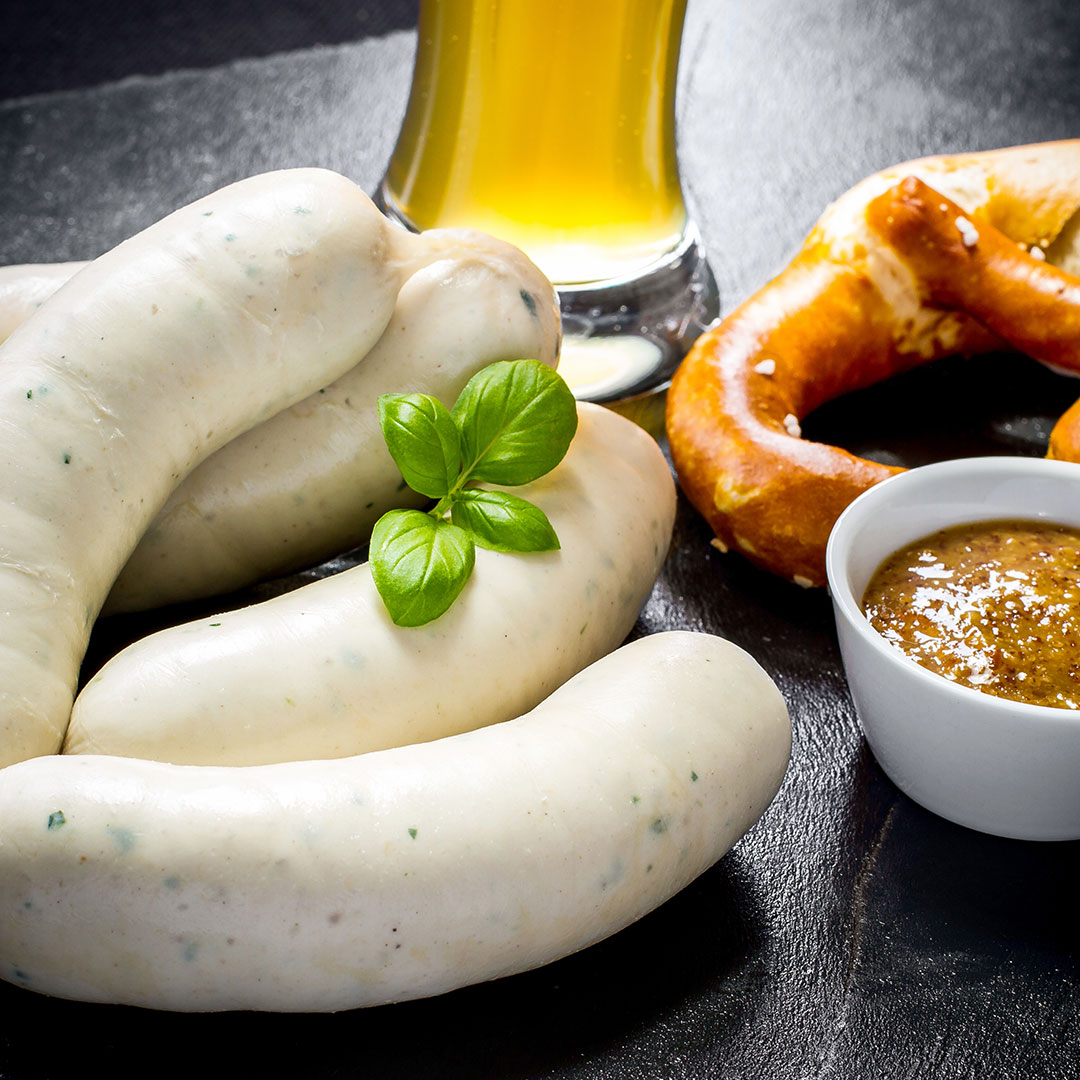 Ein Teller mit drei Weißwürsten, garniert mit Basilikum, ein Glas Bier, eine weiche Brezel und eine kleine Schale mit Senf, alles auf einer dunklen Fläche.