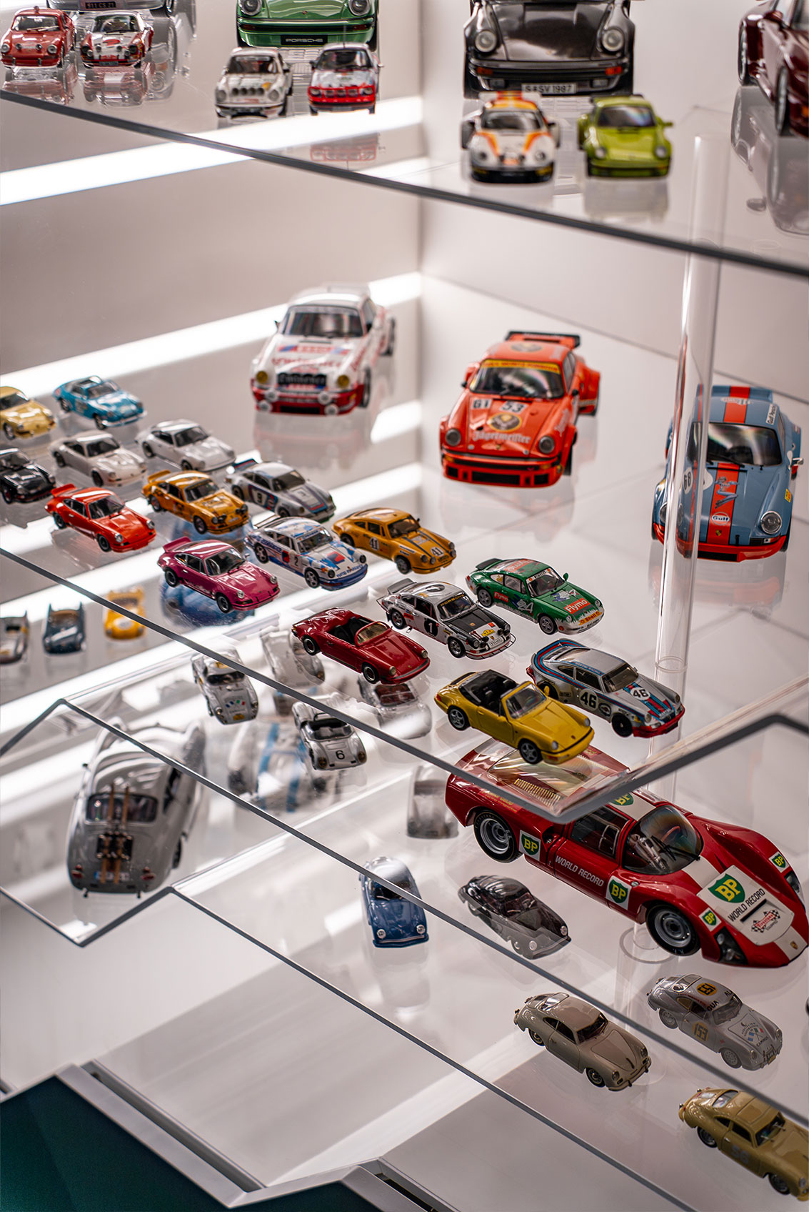 Eine Glasvitrine mit mehreren Regalen voller kleiner, detailgetreuer Modellautos in verschiedenen Farben und Stilen, die unter heller Beleuchtung fein säuberlich in Reihen angeordnet sind.