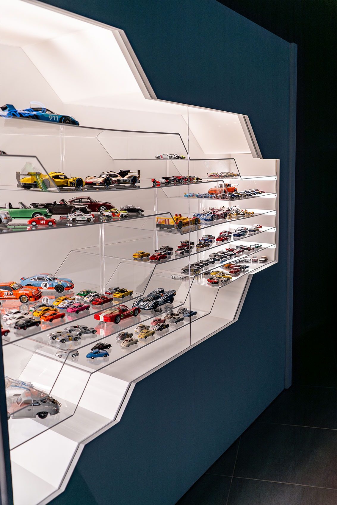 Eine moderne, eckige Glasvitrine enthält mehrere Regale mit verschiedenen Modellautos aus Diecast, darunter Rennwagen und Oldtimer, die übersichtlich angeordnet und vor einem dunklen Hintergrund hell beleuchtet sind.