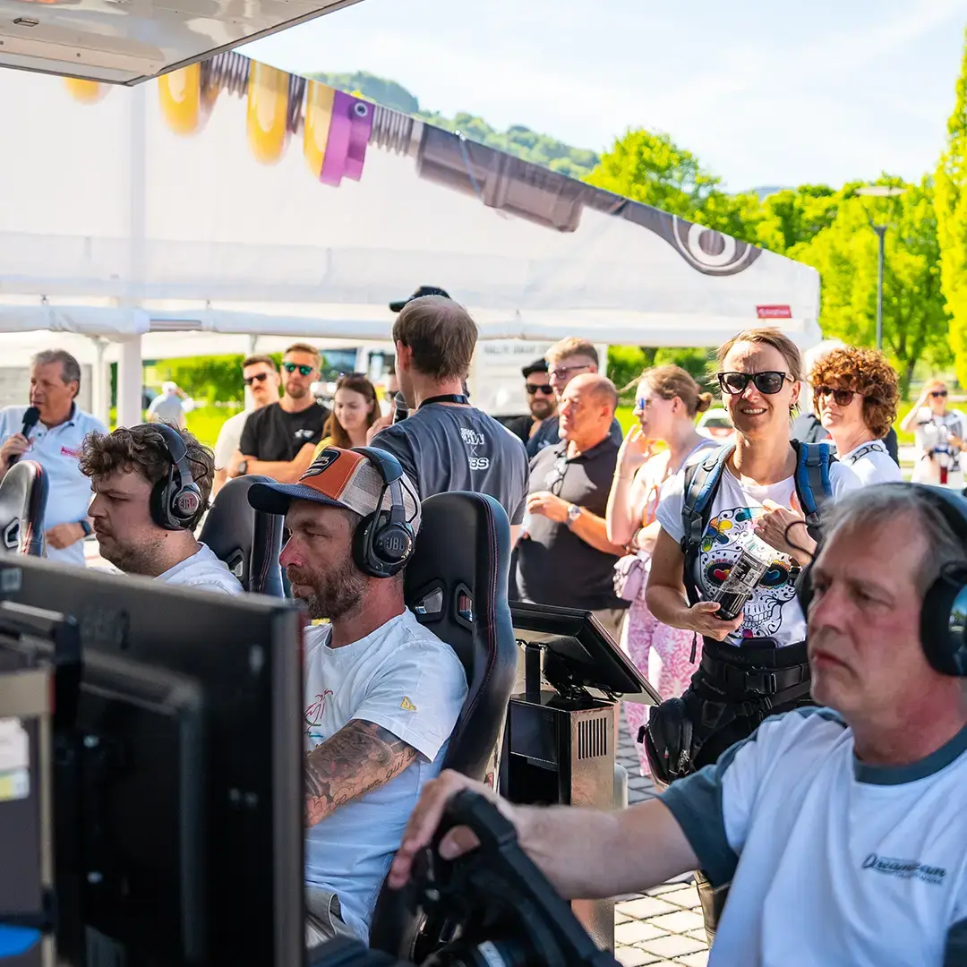 Menschen nehmen an einem Spiele-Event im Freien teil. Mehrere Personen sitzen an Computerstationen mit Kopfhörern, während andere dahinter stehen, zusehen und lächeln. Die Szene spielt sich in einem sonnigen Zelt ab, das im Hintergrund begrünt ist.