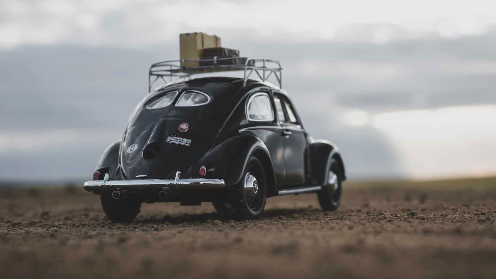 Ein schwarzer Oldtimer mit Gepäck auf dem Dachgepäckträger ist auf einem unbefestigten Weg mit einem bewölkten Himmel im Hintergrund geparkt.