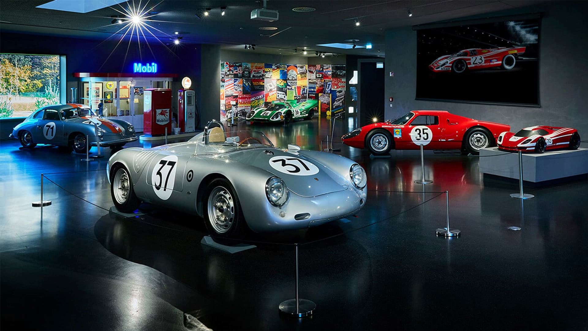 Eine Museumsausstellung mit klassischen Rennwagen, darunter ein silbernes Cabrio mit der Nummer 37 und mehrere rote Autos, die unter hellen Lichtern ausgestellt sind, mit Rennsport-Erinnerungsstücken und Fotos an den Wänden.