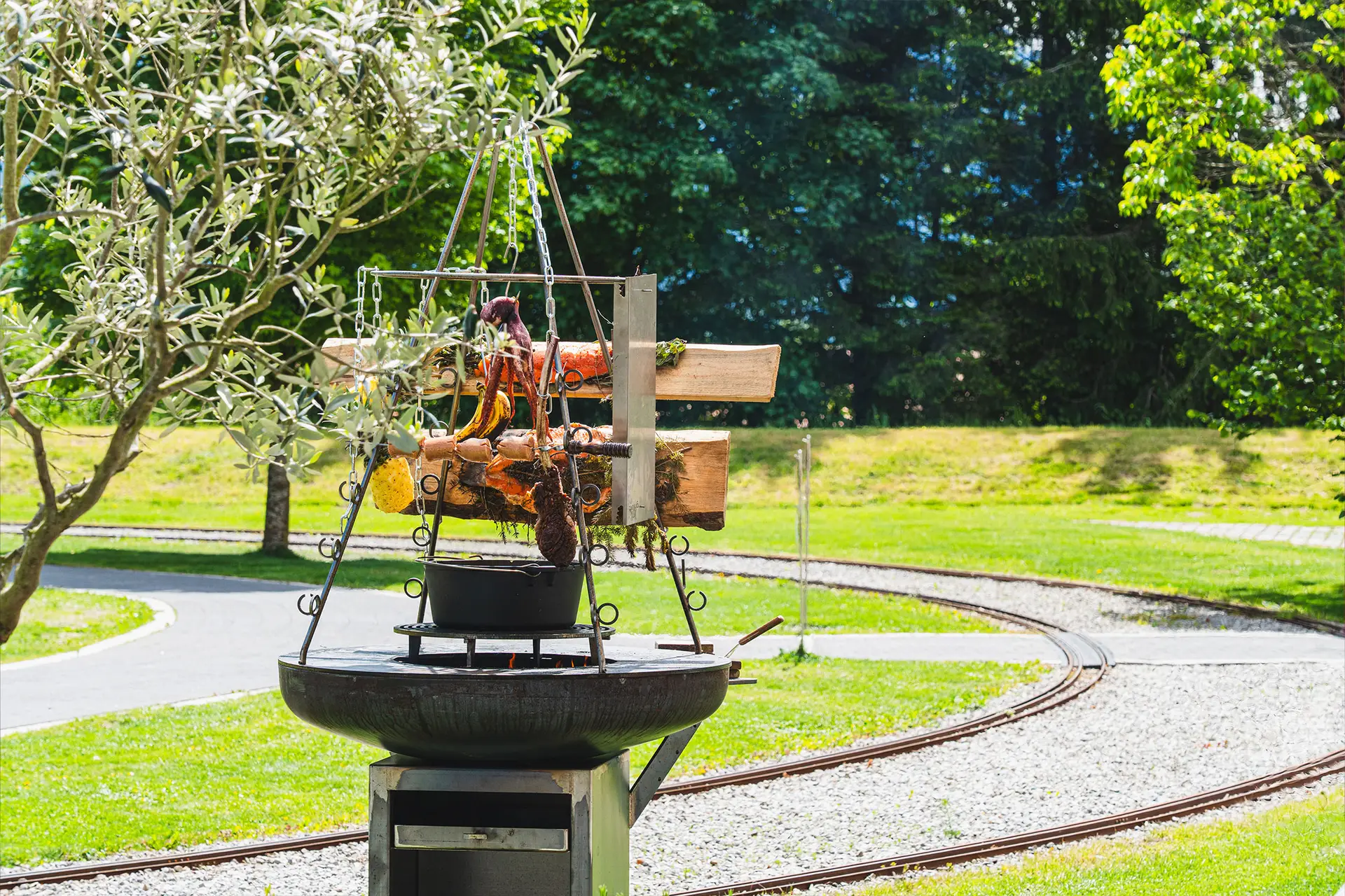 Eine dekorative Feuerstelle im Freien mit Holzbalken und Metallketten, die in der Nähe einer geschwungenen Miniaturbahnstrecke auf einer von Bäumen umgebenen Rasenfläche in strahlendem Sonnenlicht steht.
