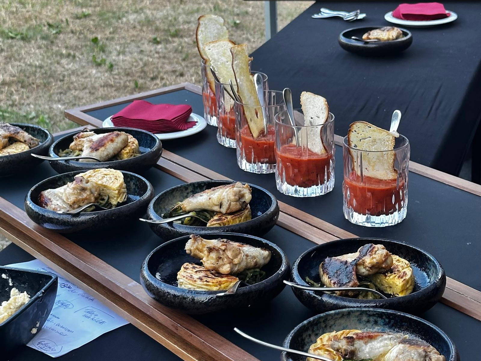 In mehreren schwarzen Schüsseln liegen gegrilltes Hähnchen und Gemüse, daneben stehen Gläser mit einer roten Suppe und Brotscheiben. Im Hintergrund stehen Teller mit roten Servietten auf den Tischen.