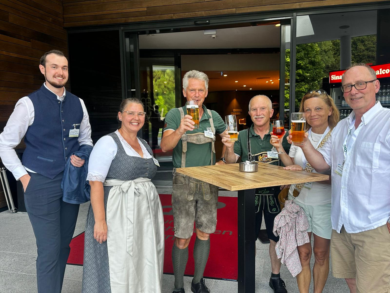 Sechs Erwachsene stehen draußen um einen hohen Tisch herum, lächeln und erheben ihre Biergläser. Einige tragen vor dem Eingang eines modernen Gebäudes traditionelle bayerische Tracht, darunter Dirndl und Lederhosen.
