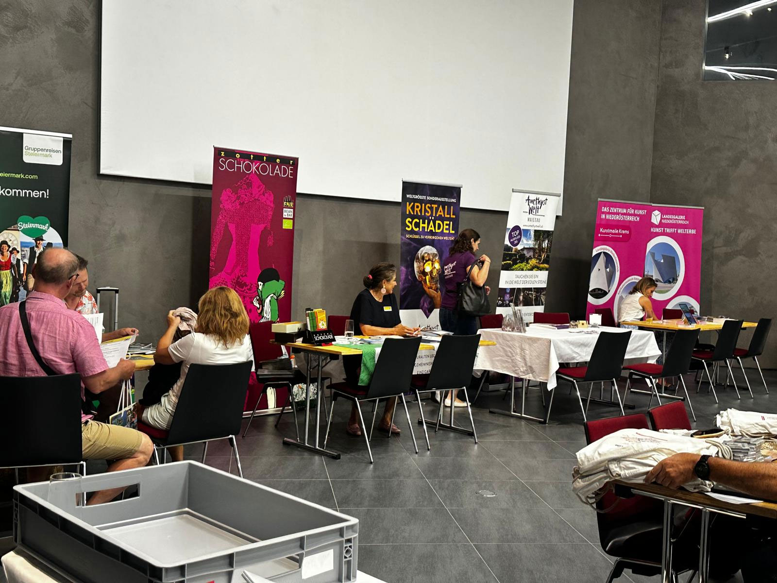 In einem modernen, grauen Raum sitzen und stehen Menschen an Tischen mit bunten Bannern, möglicherweise bei einer Veranstaltung oder Messe. Einige unterhalten sich, und auf den Tischen liegen Informationsmaterialien.
