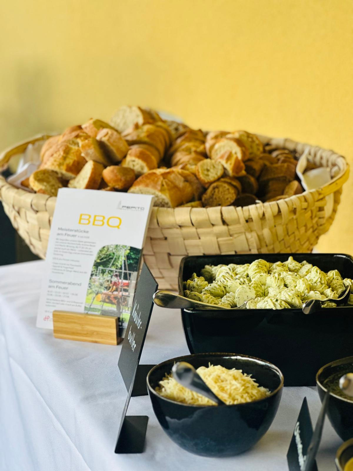 Auf einer weißen Tischdecke vor einer gelben Wand stehen ein Korb mit geschnittenem Brot, ein Schild mit der Aufschrift „BBQ“ sowie Schalen mit Aufstrich und geriebenem Käse.