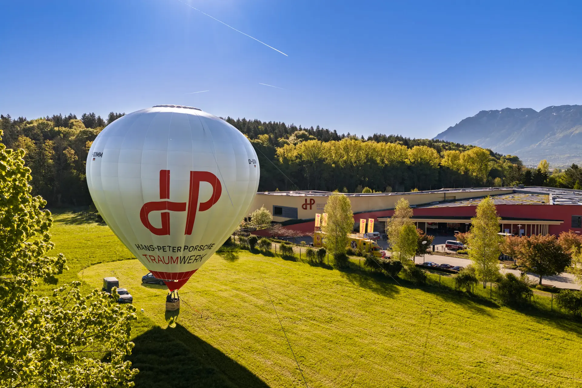Ein weißer Heißluftballon mit rotem HP-Logo und der Aufschrift „Hans-Peter Porsche Traumwerk“ wird auf einer Wiese in der Nähe eines modernen Gebäudes für den Flug vorbereitet, im Hintergrund sind Bäume und Berge zu sehen.