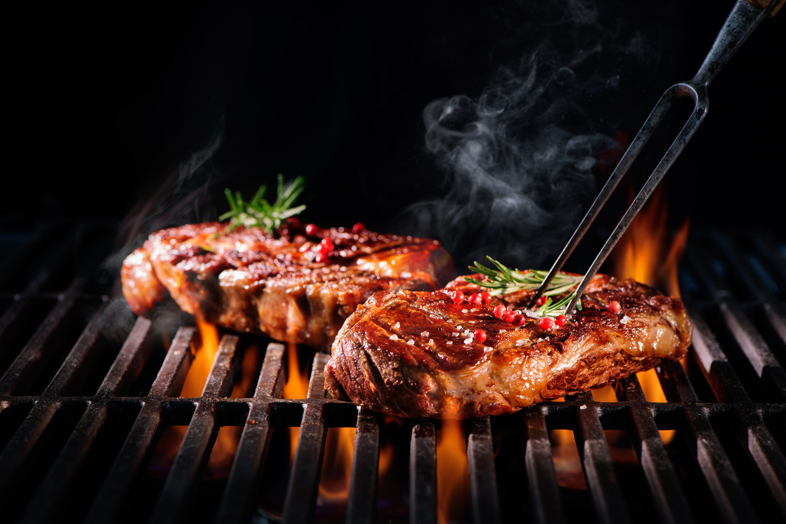Zwei saftige Steaks werden über offener Flamme gegrillt und mit Rosmarin und rosa Pfefferkörnern garniert. Eine Grillgabel hält ein Steak in die Höhe, während sich vor einem dunklen Hintergrund Rauch aufsteigt.