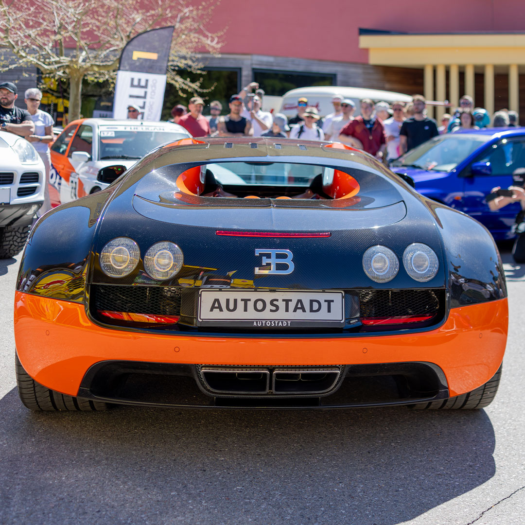 Ein schwarz-orangefarbener Bugatti-Sportwagen mit dem Kennzeichen „AUTOSTADT“ steht im Freien, umgeben von Menschen und anderen Autos auf einer Automobilausstellung.