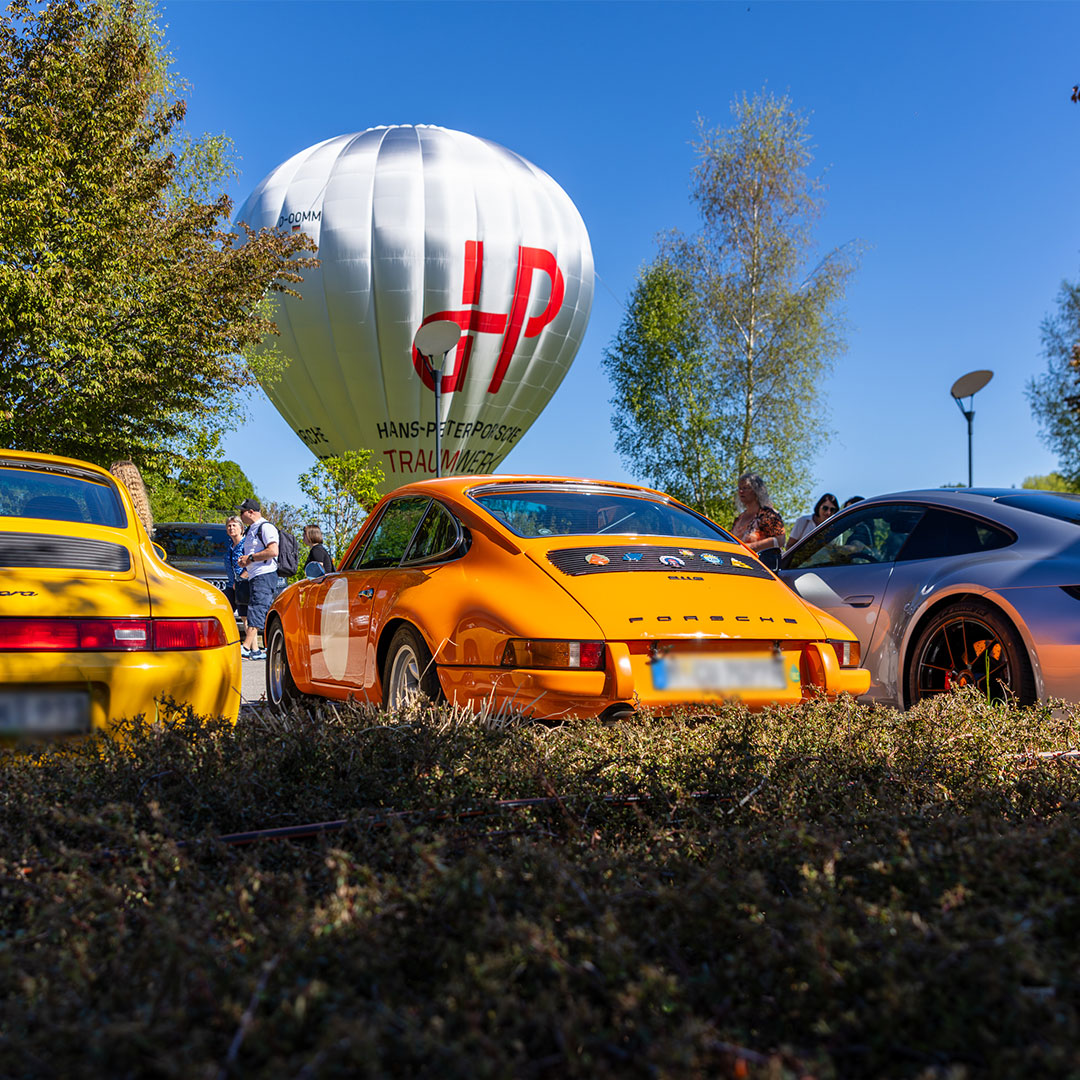 Drei Porsche-Fahrzeuge stehen im Freien in der Nähe eines großen weißen Heißluftballons mit roter Beschriftung, daneben stehen Menschen. Im Hintergrund sind Bäume und der blaue Himmel zu sehen.