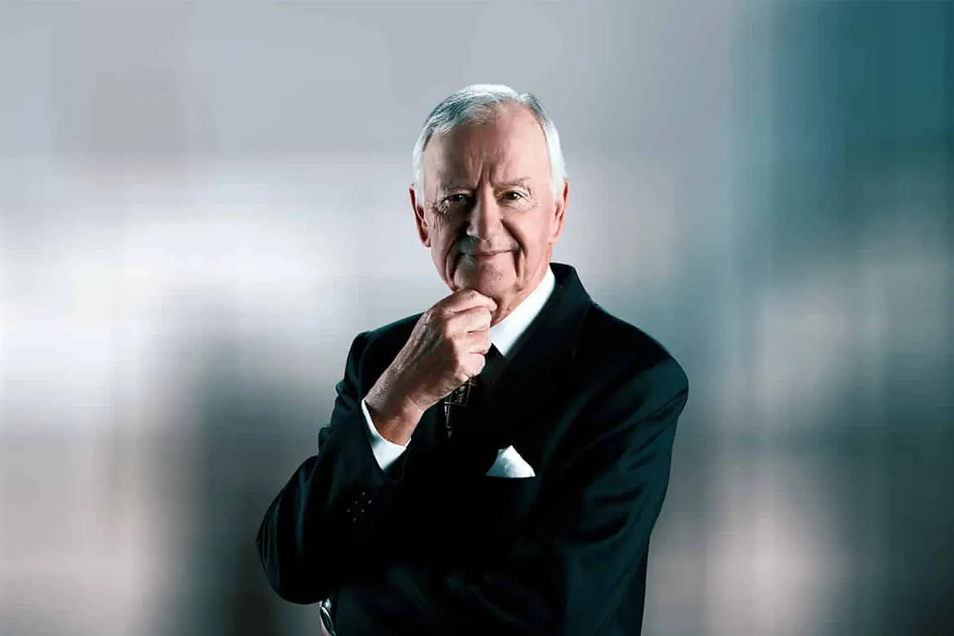 Vor einem verschwommenen blau-grauen Hintergrund steht Hans Peter Porsche in einem dunklen Anzug, der sich mit einer Hand das Kinn berührt und leicht lächelt.