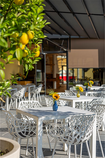 Ein Straßencafé mit weißen Tischen und kunstvoll gestalteten Stühlen, wobei jeder Tisch mit einer kleinen Vase mit Blumen geschmückt ist. Im Vordergrund hängen Zitronen an einem Baum unter einem schattenspendenden, modernen Terrassendach. Sonnenlicht taucht die Szene in helles Licht.
