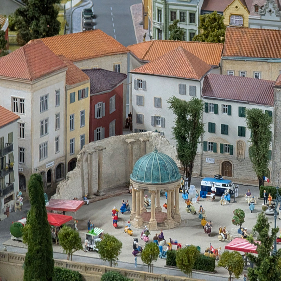 Ein detailgetreues Miniaturmodell eines europäischen Stadtplatzes zeigt farbenfrohe Gebäude, einen Pavillon mit Kuppel, Menschen, Bäume und einen blauen Polizeiwagen. Die Szene wirkt lebendig und ist äußerst detailreich gestaltet.