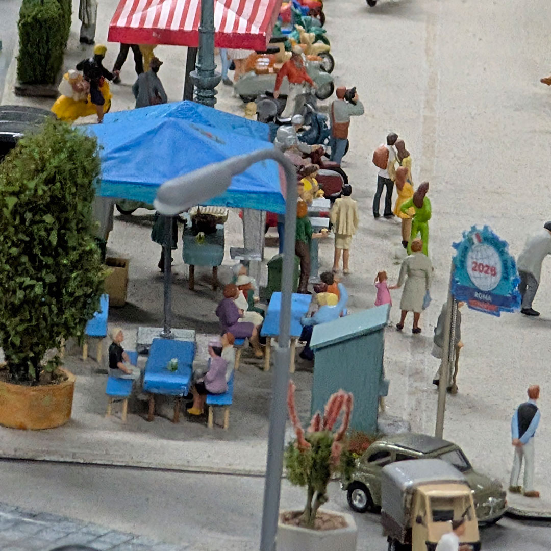Miniatur-Marktszene mit winzigen Modellfiguren an Tischen unter rot gestreiften Zelten, Laternenpfählen, Grünpflanzen und kleinen Autos, die eine lebhafte Atmosphäre im Freien einfängt. Rechts ist ein Schild mit der Aufschrift „Roma 2029“ zu sehen.