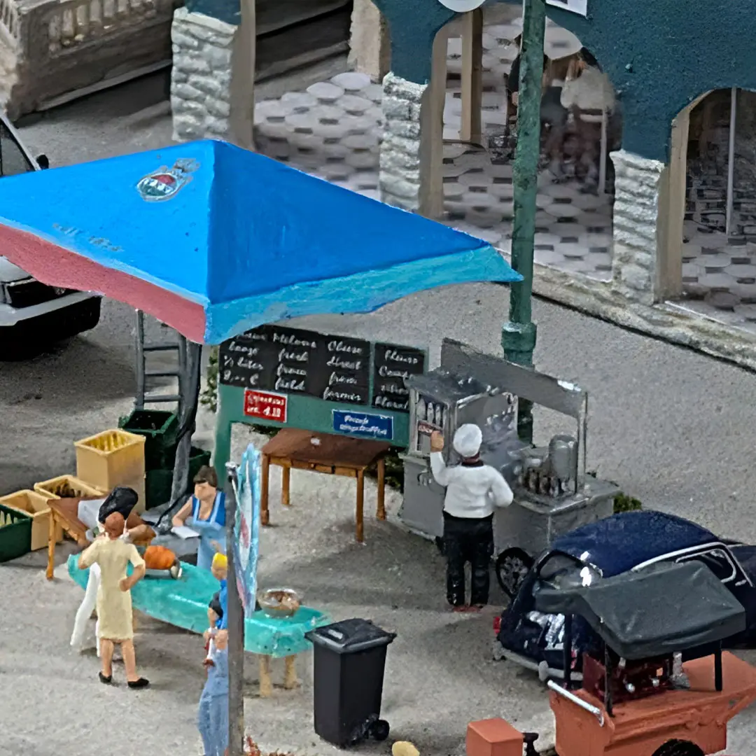 Eine Miniatur-Marktszene mit einem Verkäufer an einem Imbisswagen unter einem blauen Vordach, daneben stehenden Kunden und einem kleinen Tisch mit Lebensmitteln. Im Hintergrund sind eine Straßenlaterne und ein Gebäude zu sehen.