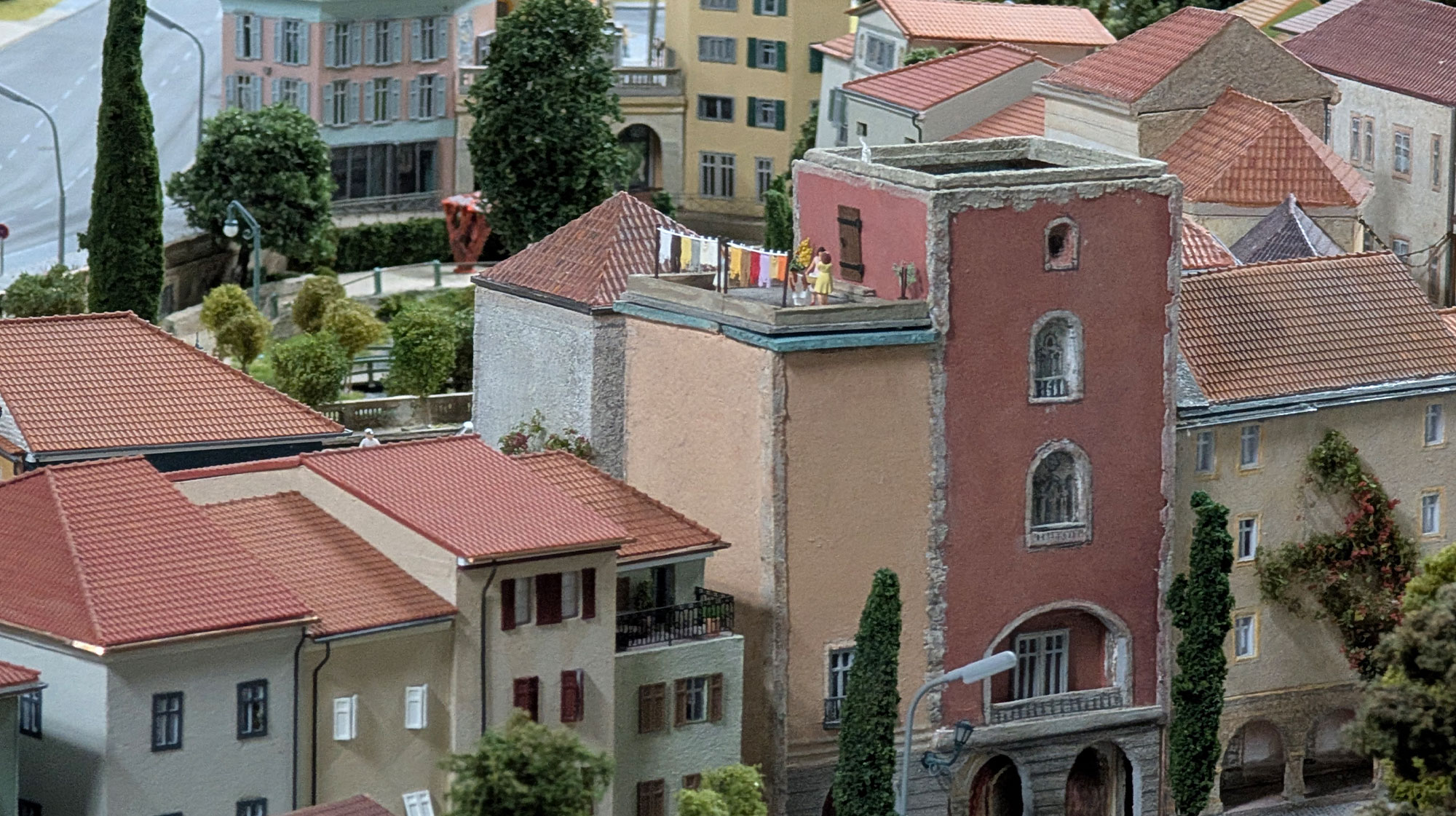Ein Modell einer Mittelmeerstadt mit bunten Gebäuden, roten Ziegeldächern und einer Dachterrasse, auf der Miniaturwäsche an einer Wäscheleine hängt. Bäume und detailreich gestaltete Fenster tragen zum realistischen Erscheinungsbild bei.