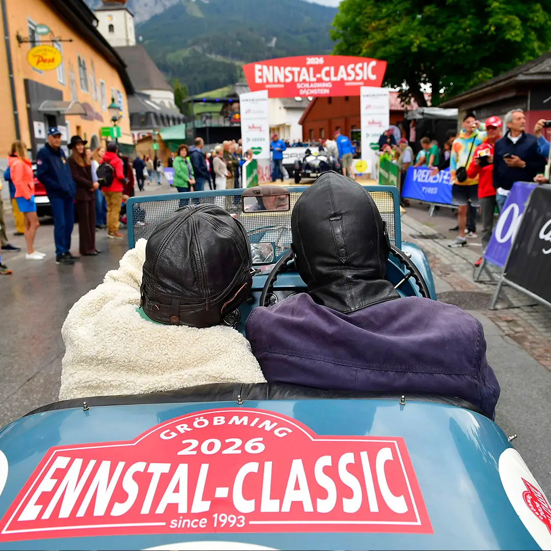 Zwei Personen in Oldtimer-Fahrerkleidung sitzen in einem Oldtimer bei der Ennstal-Classic 2026; die Aufnahme zeigt sie von hinten, während sie sich einer Menschenmenge und dem Startbanner der Veranstaltung auf einer Straße in einem Bergort nähern.