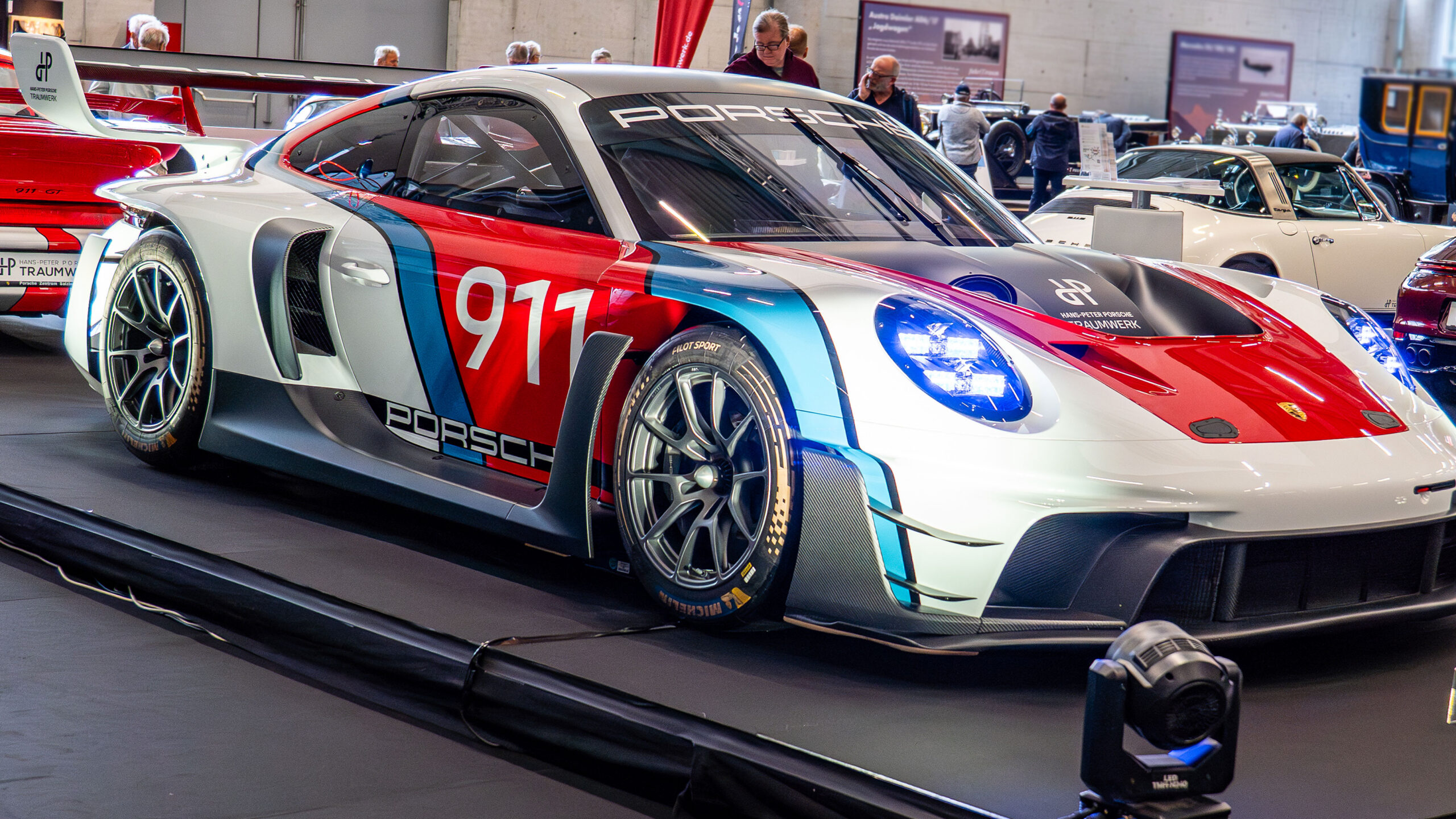 Ein Porsche-Rennwagen mit der Startnummer 911 in blau-rot-weißer Lackierung ist in den Innenräumen einer Automobilausstellung zu sehen, im Hintergrund sind weitere Oldtimer und Menschen zu erkennen.