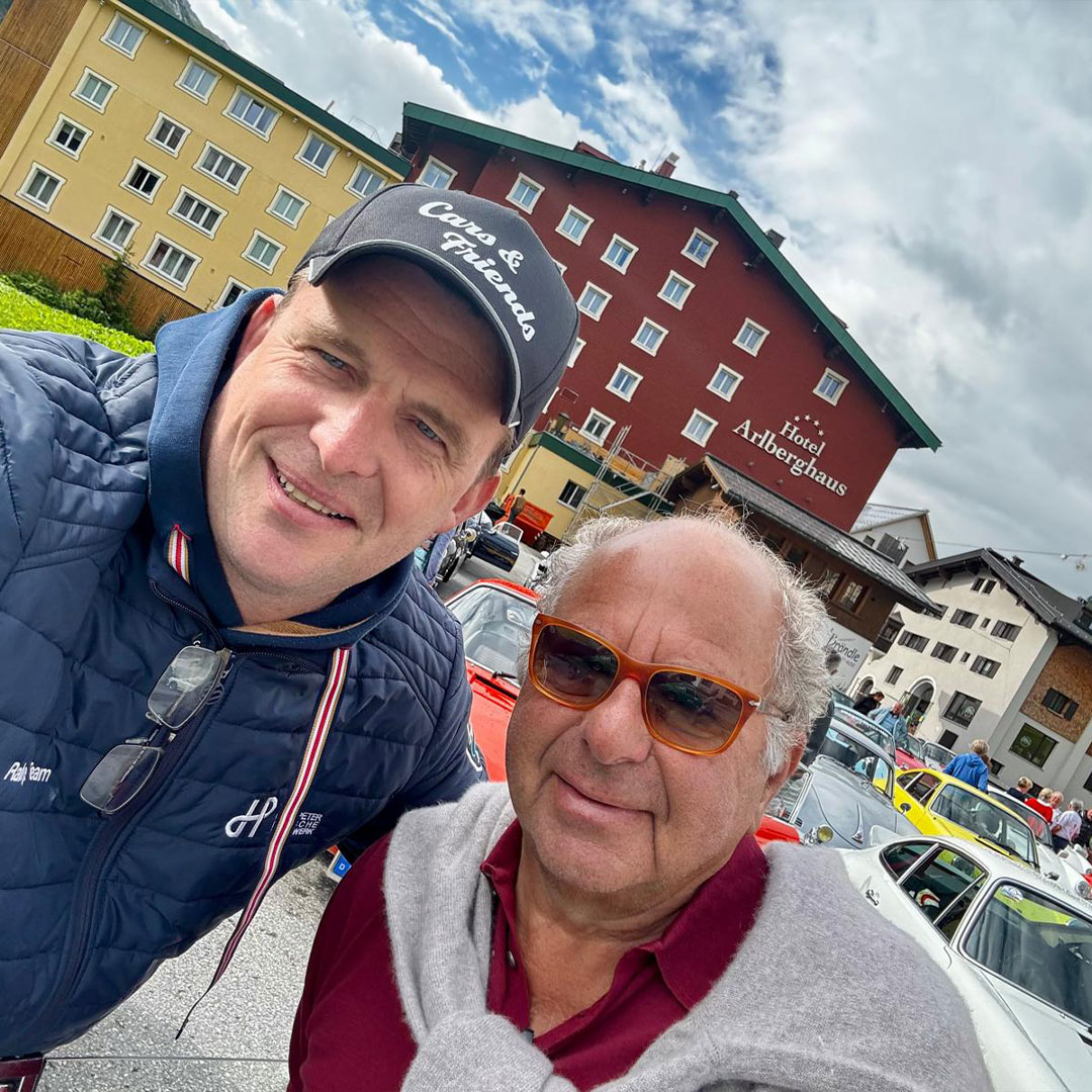 Zwei Männer lächeln im Freien auf einer Automobilausstellung, hinter ihnen stehen bunte Oldtimer. Sie stehen vor einem roten Hotelgebäude unter einem teilweise bewölkten Himmel. Der eine Mann trägt eine Sonnenbrille, der andere eine dunkle Mütze und eine blaue Jacke.