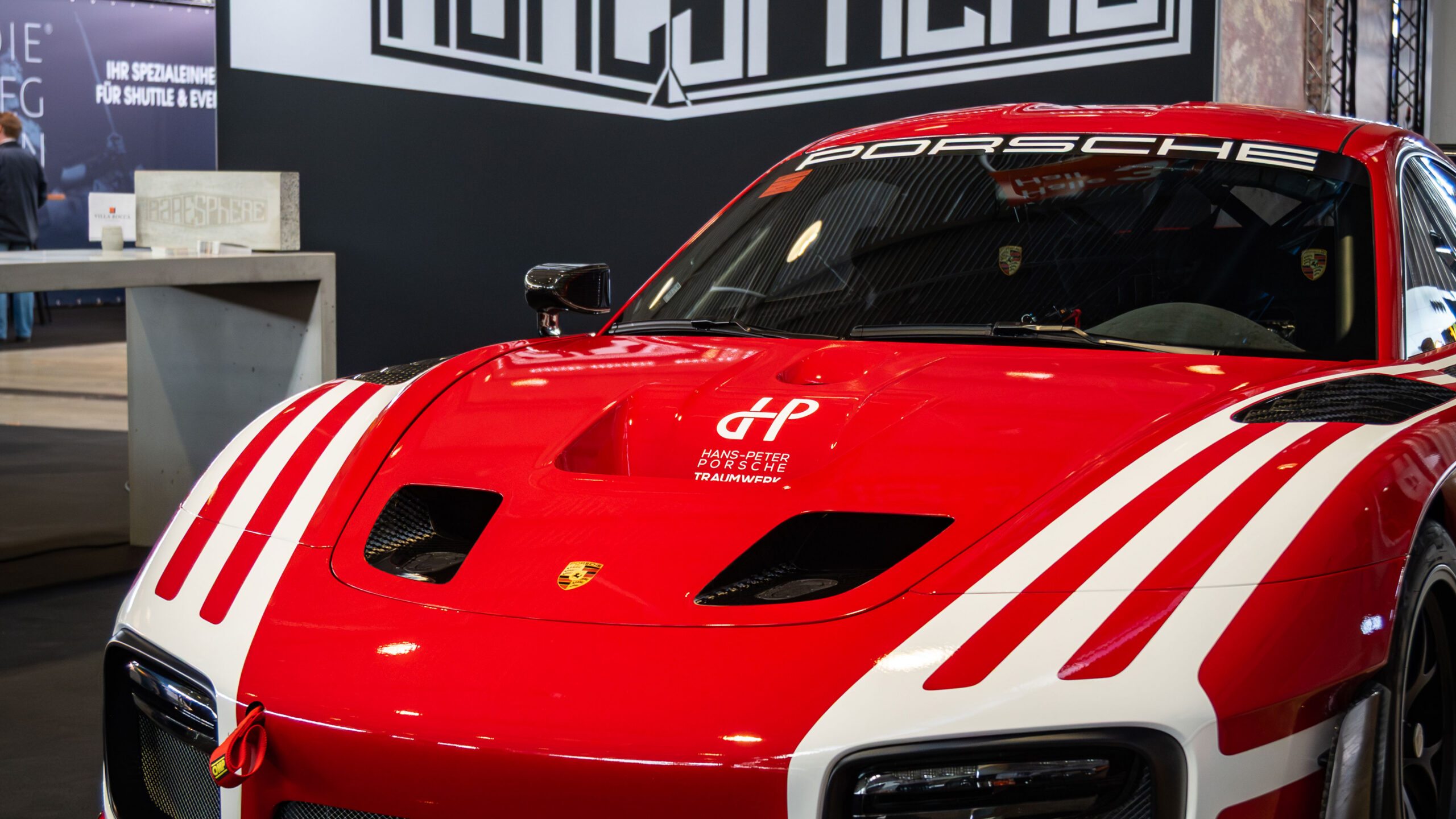 Roter und weißer Porsche-Sportwagen in einer Halle ausgestellt, Vorderansicht mit markanten Streifen und Porsche-Logo, im Hintergrund sind Beschilderungen und Tische zu sehen.