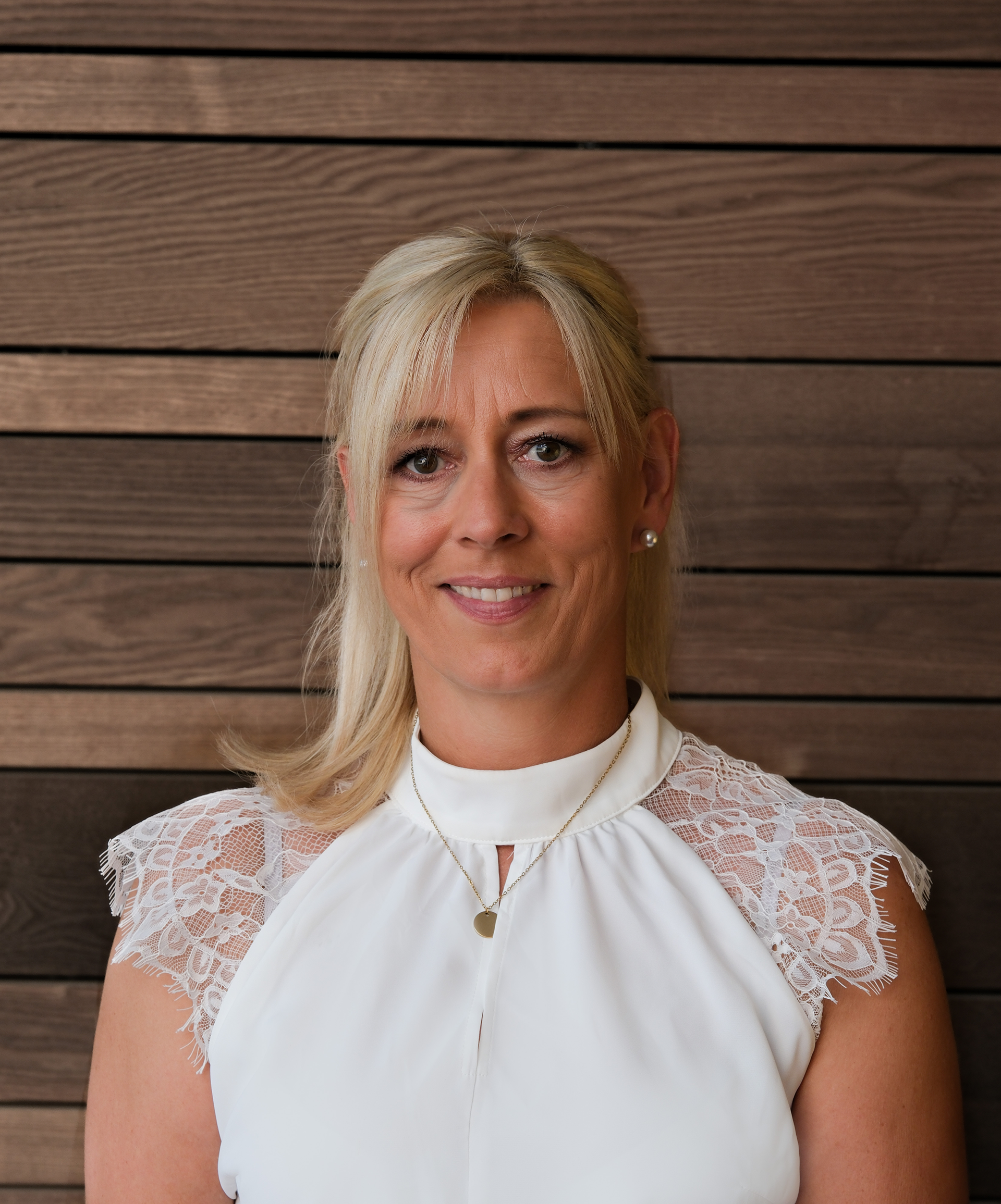 Eine lächelnde Frau mit blonden Haaren, die ein weißes Oberteil mit Spitzenärmeln und eine goldene Halskette trägt und vor einer Wand aus Holzpaneelen steht.