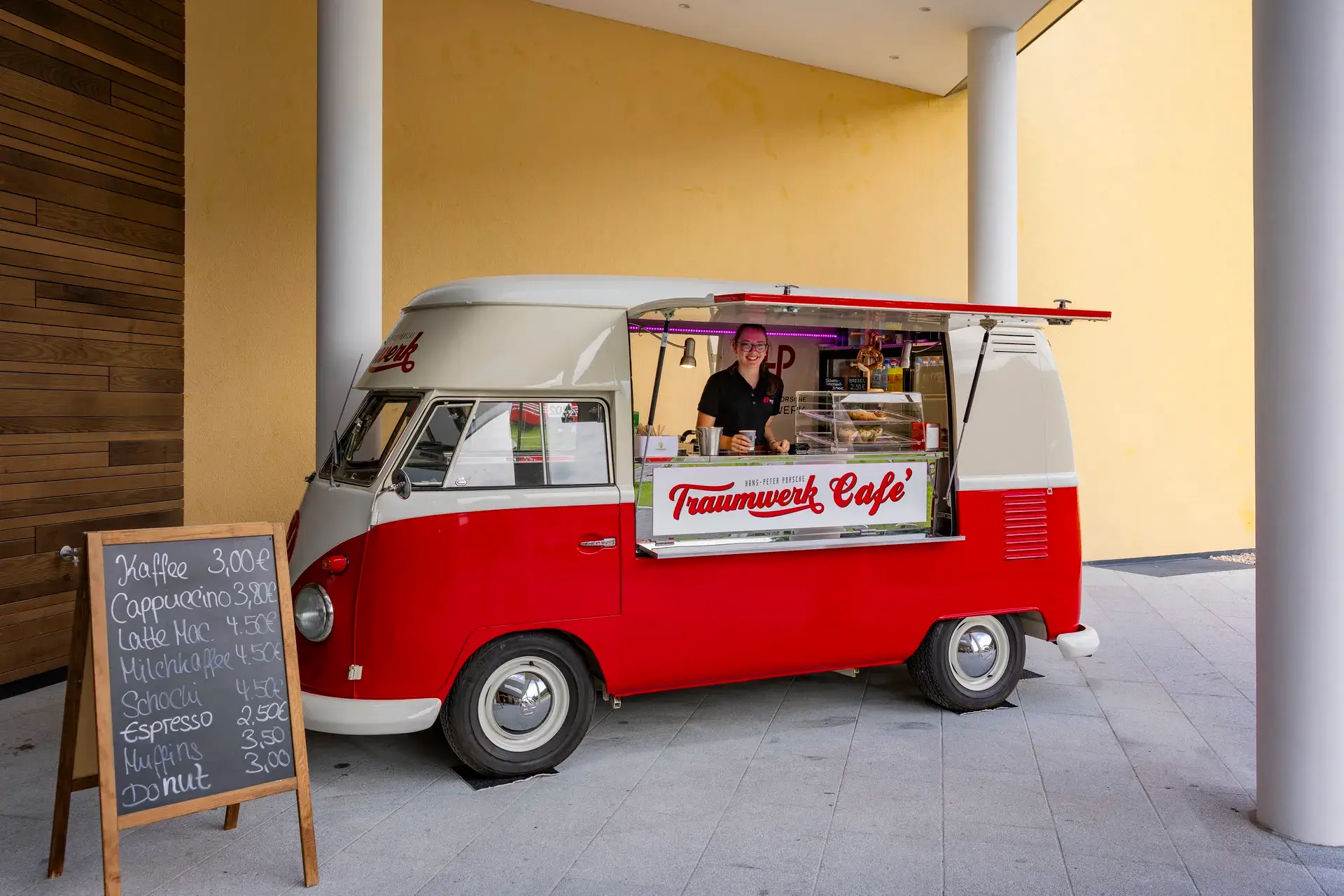 Ein rot-weißer Imbisswagen mit der Aufschrift „Traumwerk Café“ serviert Kaffee. Im Inneren steht eine Person. Draußen ist eine Kreidetafel mit der Getränkekarte und den Preisen zu sehen. Der Wagen steht in der Nähe von gelben und holzverkleideten Wänden.