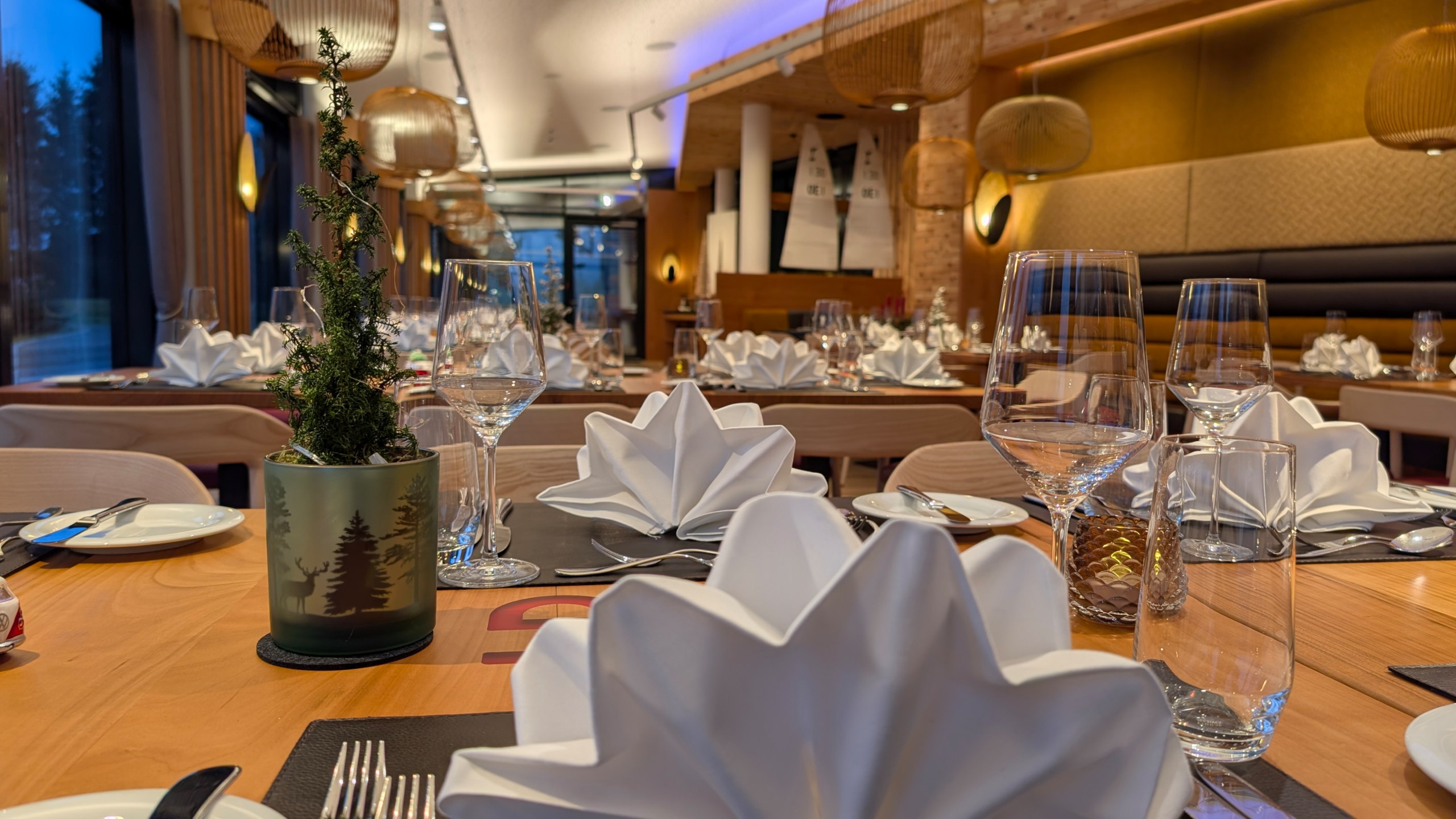 Elegantes Restaurantinterieur mit ordentlich gedeckten Tischen, gefalteten weißen Servietten, Weingläsern, Tellern, Besteck und einer kleinen Topfpflanze als Tischdekoration im Mittelpunkt. Warme Beleuchtung und modernes Dekor schaffen eine gemütliche Atmosphäre beim Essen.