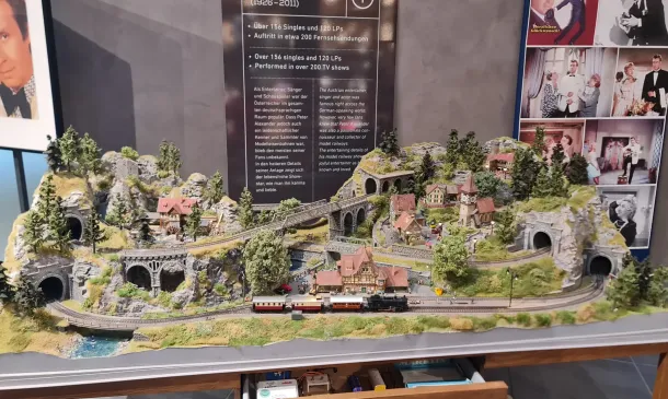 Eine detailreiche Modelleisenbahnlandschaft mit Bergen, Tunneln, Brücken, Zügen, Gleisen, kleinen Häusern und Bäumen, die in einem Innenraum ausgestellt ist, im Hintergrund sind Informationsplakate und gerahmte Fotos zu sehen.