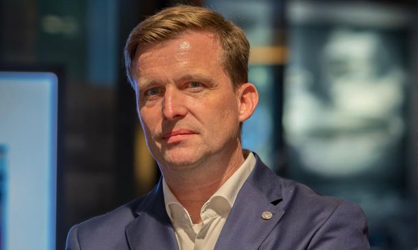 Ein Mann mittleren Alters mit kurzen hellbraunen Haaren, der eine blaue Anzugsjacke und ein weißes Hemd trägt, steht in einem Haus und hat einen neutralen Gesichtsausdruck. Der Hintergrund ist unscharf.