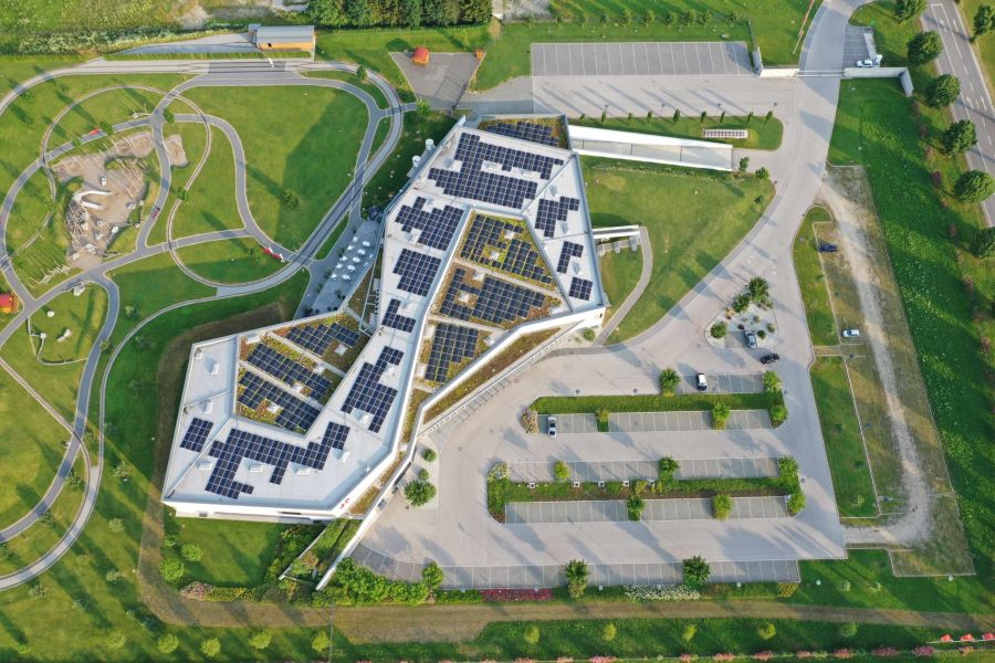 Luftaufnahme eines modernen, eckigen Gebäudes mit einem mit Solarmodulen bedeckten Dach, umgeben von grünen Rasenflächen, einem Spielplatz, Parkplätzen, Bäumen und Gehwegen.