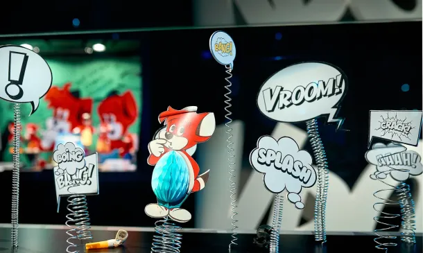 Comic-Display mit einem Zeichentricktier, Sprechblasen mit den Worten „VROOM!“ und „SPLASH!“ sowie an Schildern befestigten Federn. Auf der Oberfläche liegt ein gelbes Horn, im Hintergrund ist buntes Zeichentrick-Design zu sehen.