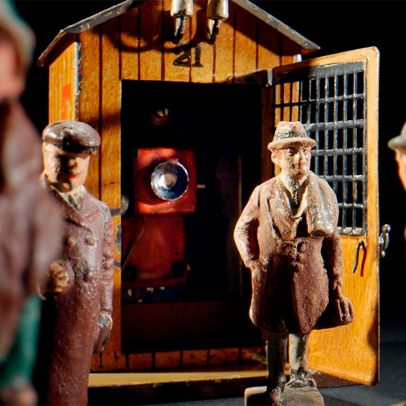 Mehrere kleine, antike Figuren stehen vor einem Holzschuppen mit offener Tür, in dessen Inneren sich eine altmodische Kamera befindet. Die Szene ist schwach beleuchtet und schafft eine dramatische, nostalgische Atmosphäre.